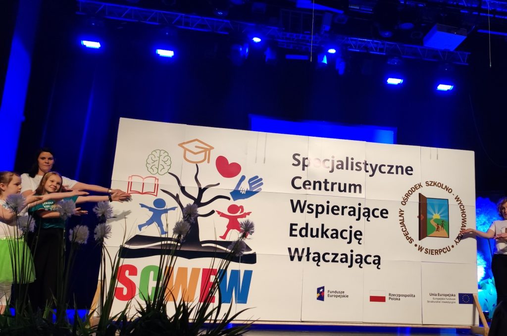 Zdjęcie przedstawia dzieci wskazujące na plakat, na którym jest napisane Specjalistyczne Centrum Wspierające Edukację Włączającą