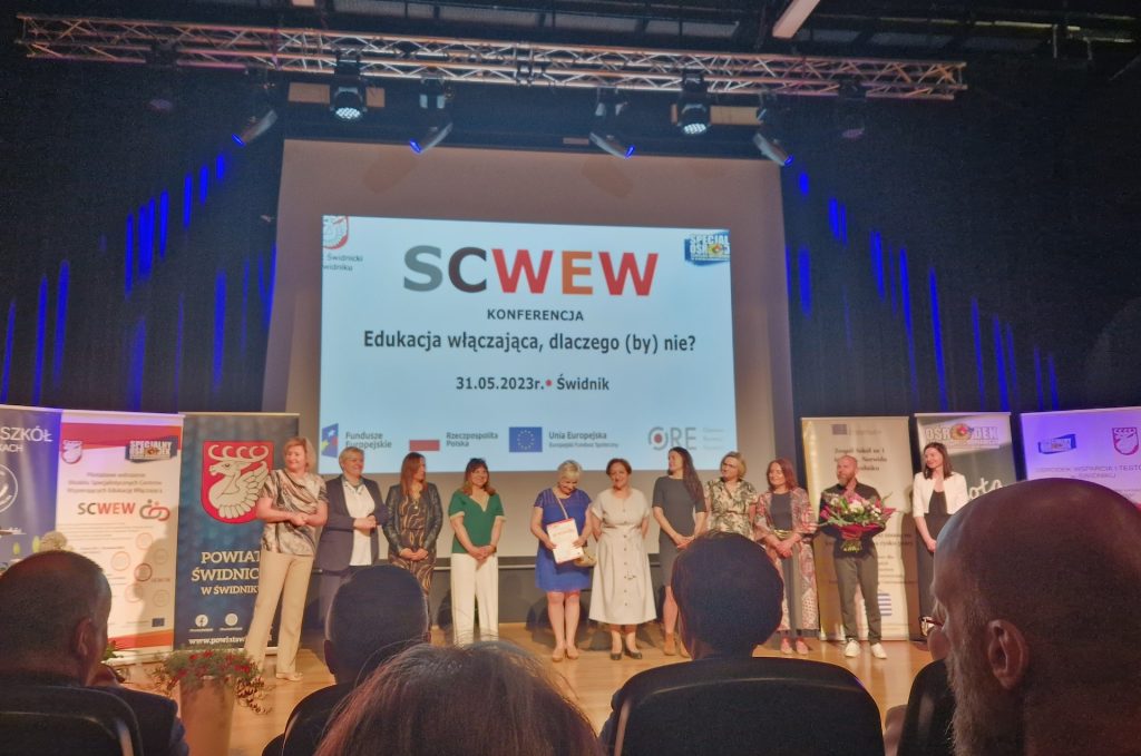 Zdjęcie przedstawia scenę, na której w środkowej części stoi 11 osób – koordynatorów i ekspertów SCWEW. W lewej części zdjęcia widać Panią Elżbietę Krupę – Lidera SCWEW, stojącą przy mównicy z mikrofonem. Za ludźmi stojącymi na scenie stoją roll-upy oraz wisi ekran, na którym wyświetlany jest slajd
