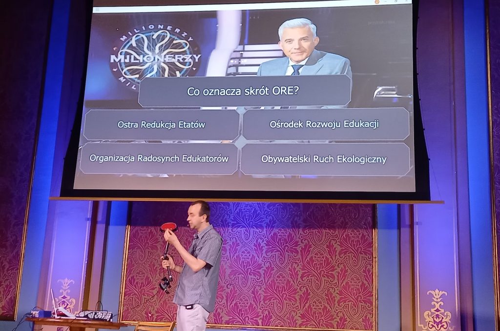 Zdjęcie przedstawia osobę, która trzyma w rękach sprzęt dla osób, które mają problem z rękoma na tle dużego ekranu