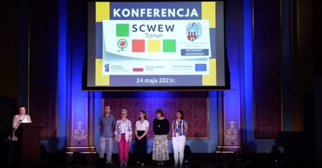 Zdjęcie przedstawia cztery osoby na tle dużego ekranu, na którym jest napis SCWEW i przy mikrofonie z lewej strony stoi jedna osoba.