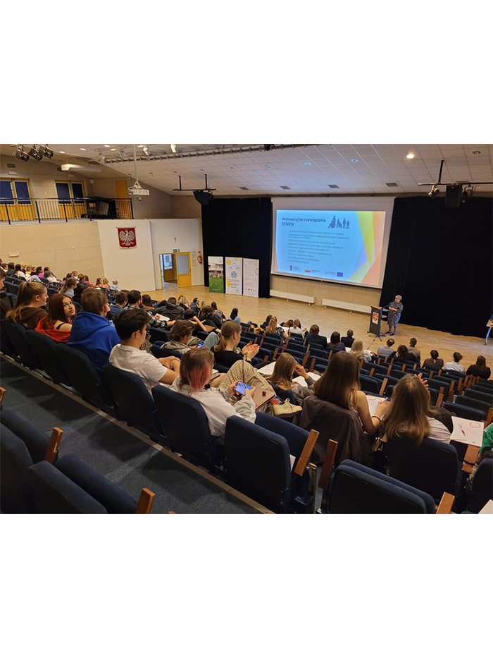 Prelekcja Kierownik projektu z Ośrodka Rozwoju Edukacji podczas konferencji w Łomiankach. Na ekranie wyświetlona jest prezentacja. Publiczność siedzi na krzesłach.