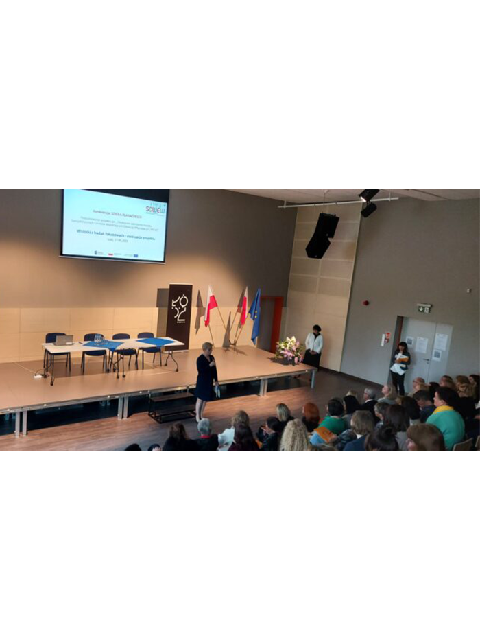 SCWEW Łódź - konferencja. Widok dużej sali. Z przodu sali ekran i monitor. Do mikrofonu mówi kobieta. Publiczność siedzi na krzesłach.