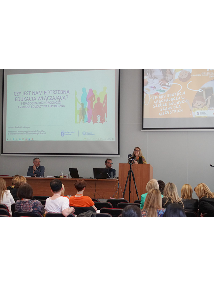 Zdjęcie przedstawia salę konferencyjną. W górnej części zdjęcia widoczne są dwa ekrany z prezentacją. W środkowej części zdjęcia znajduje się długi stół prezydialny przy którym siedzą dwie osoby, z prawej strony stołu stoi mównica, na której przemawia prelegentka. Na dole zdjęcia widoczne są krzesła i siedząca widownia.