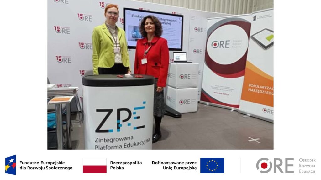 Przedstawicielki zespołu projektu Katarzyna Lidia Kuklińska i Barbara Wałecka stoją przy stole. Na stole leżą ulotki Ośrodka Rozwoju Edukacji i ulotki projektu. W tle znajdują się stojak reklamowy z napisem „ORE”, monitor z wyświetlanym filmem promocyjnym i ścianka z napisem „15-lecie ORE”