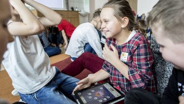 Dzieci siedzą na podłodze w sali szkolnej. Dziewczynka na pierwszy planie trzyma na kolanach tablet.