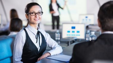 W pomieszczeniu znajdują się cztery osoby. Na pierwszym planie odwrócony plecami mężczyzna rozmawiający z uśmiechniętą kobietą. W tle stoją dwie osoby, ich postaci są rozmyte