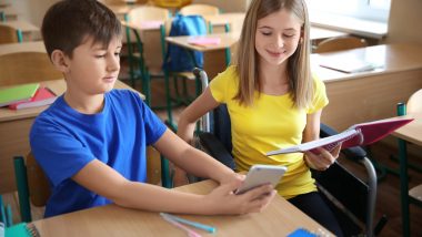 Klasa szkolna. Za jedną ławką siedzi chłopiec w niebieskiej bluzce. Obok dziewczynka w żółtej bluzce na wózku inwalidzkim. Uczniowie pracują: dziewczynka ma w ręku notatnik, a chłopiec smartfon. Na ławce leżą dwa długopisy.