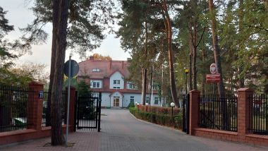 Otwarta brama wjazdowa na końcu której widać biały pałacyk z czerwonym dachem.