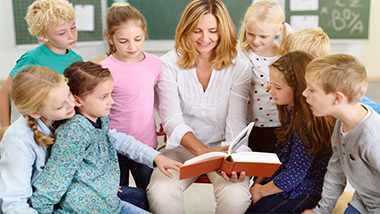 Zdjęcie, które przedstawia kobietę trzymającą książkę w dłoniach w otoczeniu dzieci