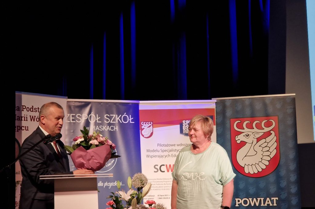 Zdjęcie przedstawia dwie postacie: po lewej stronie stoi Pan Romuald Gumienniak, który trzyma w rękach bukiet kwiatów, zaś po prawej stronie stoi Pani Elżbieta Krupa, Lider SCWEW. Za plecami tych osób stoją cztery roll-upy.