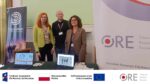 Przedstawiciele zespołu projektu: Katarzyna Lidia Kuklińska, Sebastian Dmochowski i Barbara Wałecka stoją przy stole. Na stole znajdują się ulotki Ośrodka Rozwoju Edukacji i ulotki projektu oraz dwa otwarte laptopy. Z prawej strony stołu stoi stojak reklamowy z napisem "ORE".